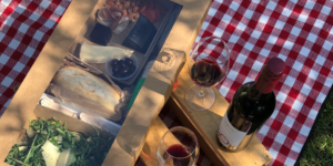 a brown card box with picnic food, on a wooden table with a bottle of wine and two glasses